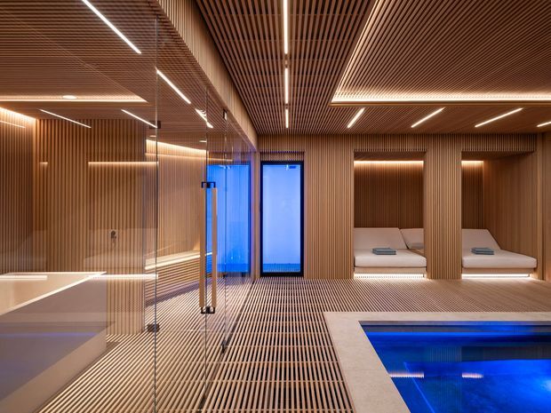 A refined spa and pool interior clad extensively in warm timber slats. The image shows a blue-lit plunge pool in the foreground, glass sauna or steam-room doors, built-in daybeds, linear integrated lighting, and a slatted timber ceiling and floor creating a highly coordinated wellness environment.