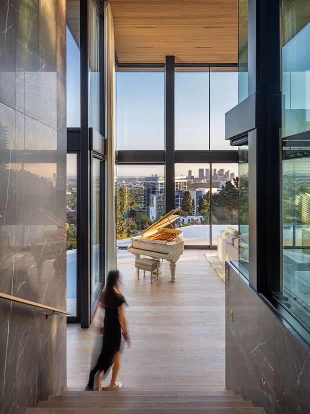 A dramatic double-height interior stair hall opening into a bright living space with floor-to-ceiling glazing and expansive city views. Dark marble-clad walls and stair surfaces contrast with a warm timber-lined ceiling, pale timber flooring, black structural framing, and a white grand piano. A person is visible on the stairs, while reflective glazing and polished surfaces add depth.