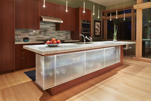 A warm, contemporary kitchen centred on a large island with a pale stone benchtop and brushed translucent metal-look cabinetry panels. The kitchen features rich reddish-brown wood cabinetry, a horizontally patterned grey tiled splashback, integrated stainless-steel appliances, pendant lighting, and light timber flooring. A glazed timber-framed opening and dining area are visible beyond the kitchen.