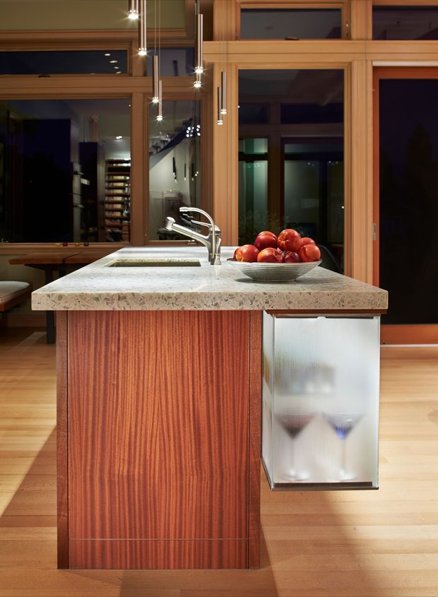 A warmly lit contemporary kitchen focused on a compact island. The island features a heavily patterned pale stone benchtop with an integrated sink and curved chrome tap, vertically grained reddish timber cabinetry, and a translucent frosted-glass side panel. Timber-framed floor-to-ceiling glazing and dark outdoor views form the backdrop, with slim pendant lights above and timber flooring throughout.