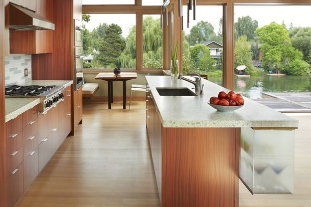 A bright contemporary kitchen with warm vertical timber cabinetry, a large pale stone island with integrated sink and seating, stainless-steel appliances, and expansive timber-framed glazing overlooking a leafy waterside setting. A small dining nook sits beside the windows.