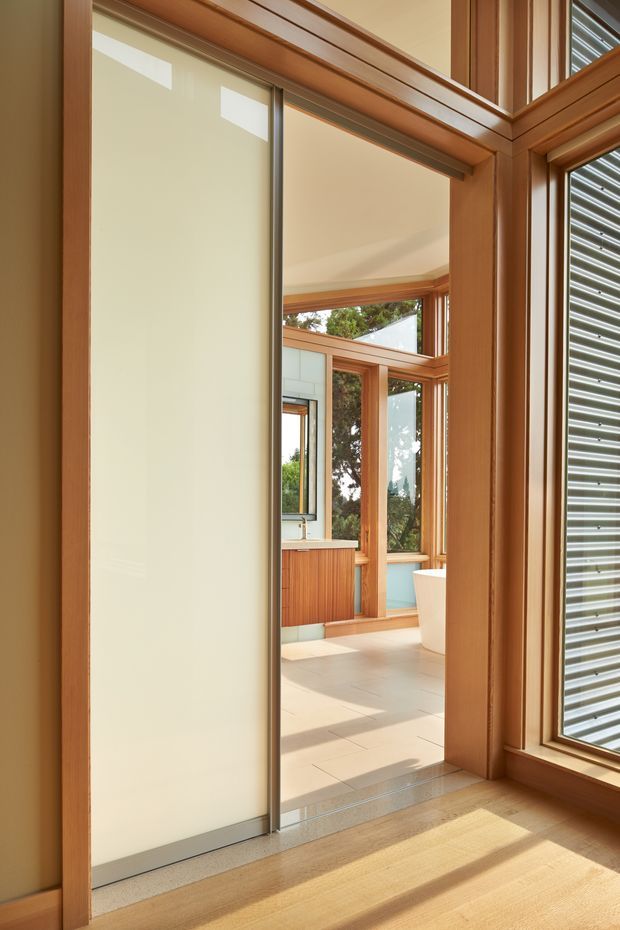 A warm, light-filled interior view through a timber-framed opening into a bathroom. The foreground has a frosted-glass sliding door, honey-toned timber flooring and substantial timber trim. Beyond it are pale tiled floors, timber-framed glazing, a timber vanity with a pale benchtop, and part of a freestanding white bathtub. Exterior greenery is visible through the windows.