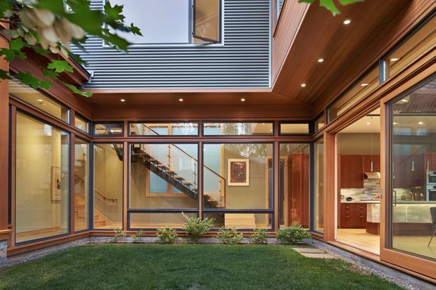 A contemporary courtyard viewed from within, enclosed by warm timber-clad walls and extensive dark-framed glazing. The courtyard has a lawn, gravel borders, and low planting, while the glazed interior reveals a stairway, hallway, and kitchen. A large overhead opening with horizontal metal cladding and roof glazing brings light into the space.