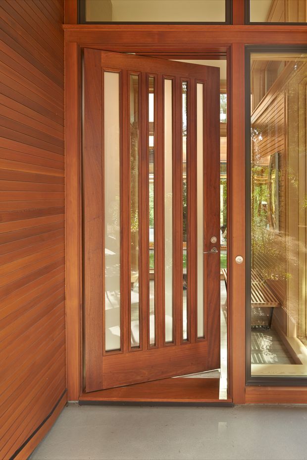 Close-up view of a contemporary timber-framed entry door with multiple narrow vertical glazed panels, set within warm reddish-brown horizontal timber wall cladding. The door is open, revealing a bright interior and a glazed side panel with reflections of the surrounding timber architecture. A smooth grey concrete floor forms the threshold.