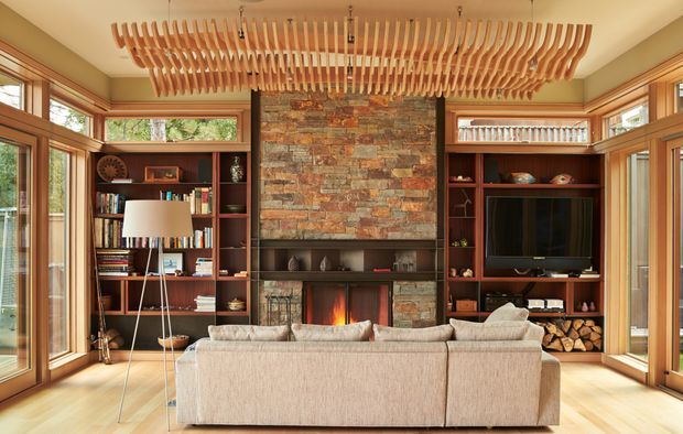 A warm, timber-framed living room with extensive built-in dark wood shelving, a central natural stone fireplace, and a large textured wood-slat pendant light. Floor-to-ceiling glazing and corner windows bring in views of greenery, while a neutral sectional sofa faces the fireplace and television.
