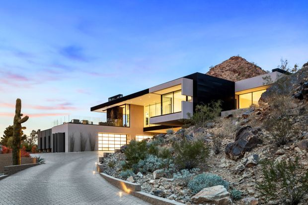 A modern multi-level hillside residence at dusk, set into a rocky desert landscape. The home features a flat roof, strong cantilevered forms, expansive floor-to-ceiling glazing, warm interior lighting, dark and light facade volumes, and an integrated garage. A paved driveway curves toward the house, with xeriscaped planting and a tall cactus in the foreground.