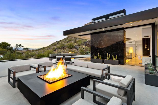 A contemporary rooftop terrace or balcony lounge adjoining a modern home, with a large black rectangular fire table, pale upholstered outdoor sofas and armchairs, and a dramatic dark stone feature wall with built-in planters. The terrace has a low glass balustrade overlooking a rocky hillside landscape, while the open interior beyond shows warm timber flooring, recessed lighting, and minimalist furnishings. The flat-roofed building has deep projecting eaves and rooftop screening elements.