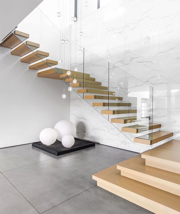 A minimalist contemporary stairway with floating light timber treads, slim glass balustrades, and a dramatic multi-level stair configuration. Large white marble-look veined wall and ceiling surfaces form the backdrop, while spherical pendant lights hang at varying heights above a dark plinth with white decorative spheres. The space is bright, refined, and highly geometric.