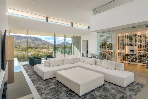 A bright, spacious contemporary living room with a large white modular sectional sofa and oversized ottoman on a grey rug. Floor-to-ceiling glazing provides expansive views of a mountainous landscape, while a glass-enclosed wine display room is visible to the right. The space has white walls, a high ceiling with an upper-level overlook, integrated lighting, and a sleek, luxurious open-plan arrangement.