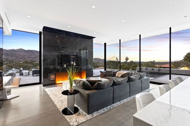Open-plan luxury living and dining area with a large grey sectional sofa arranged around a black marble fireplace with an integrated linear fire. Floor-to-ceiling glazing surrounds the room and opens to expansive mountain and suburban views. A white marble dining table with upholstered white chairs occupies the foreground, while polished concrete flooring, dark slender window frames, and a neutral contemporary palette create a refined, airy interior.