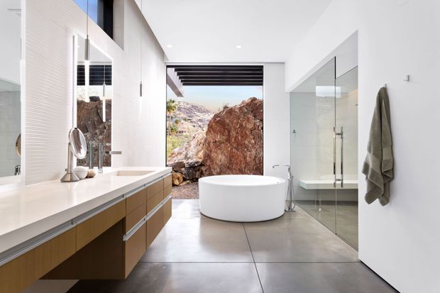 A bright, contemporary bathroom with a long floating timber vanity and white benchtop, large-format grey concrete-look floor tiles, a freestanding white oval bathtub, and a frameless glass shower enclosure. A full-height opening frames rugged natural rock outside, while a large mirror, white walls, and dark horizontal ceiling battens add architectural interest. The space feels clean, spacious, and strongly connected to its rocky landscape setting.