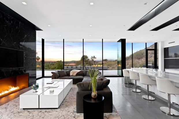 A luxurious open-plan living area with a dark marble feature wall, built-in fireplace and television, large sectional sofa, white geometric coffee table, and floor-to-ceiling glazing. The adjoining kitchen or bar area has a long white island with integrated seating. The room is bright and contemporary, with expansive views across the landscape.