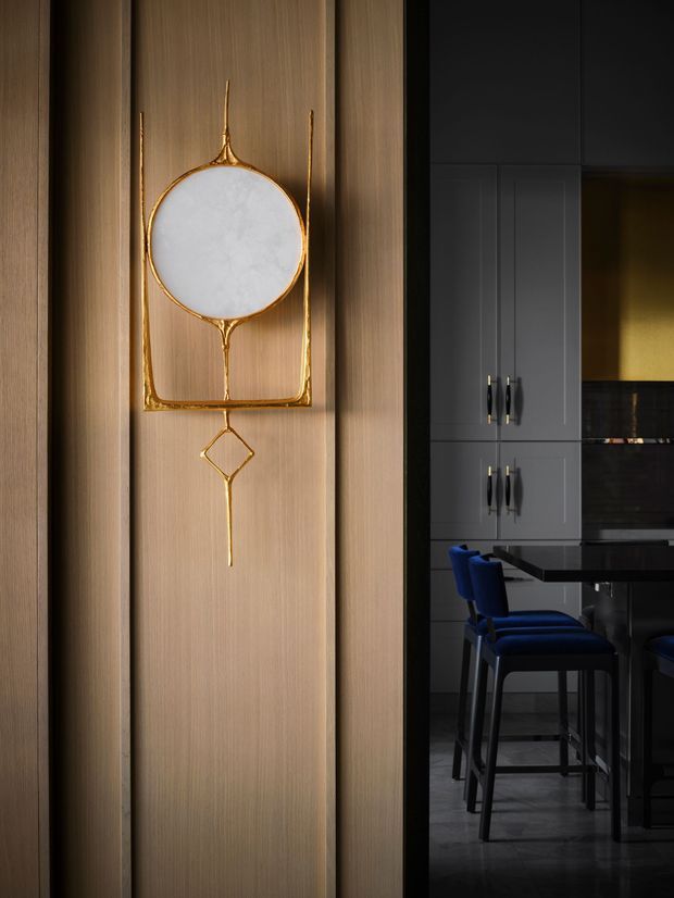 A moody, close-up interior view of a sophisticated kitchen. The foreground features tall vertical timber wall panelling with a sculptural circular gold wall ornament and white inset detail. Through the dark-framed opening, a contemporary kitchen is visible with charcoal-grey cabinetry, a dark island or counter, black reflective splashback surfaces, and deep blue bar stools.