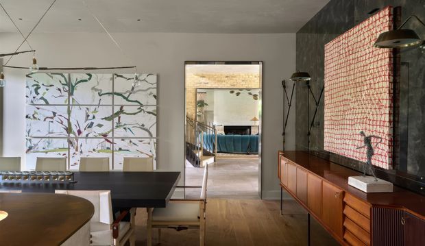 Interior view of a refined open-plan dining and living area. A dark rectangular dining table with upholstered chairs occupies the foreground, while a long mid-century-style timber sideboard runs along a dark feature wall. The wall includes a large red-and-white patterned artwork or textile panel, sculptural lighting, and a framed mirror or opening reflecting a blue sofa and staircase beyond. Light timber flooring, a pale ceiling, and restrained contemporary furnishings create a warm, curated atmosphere.