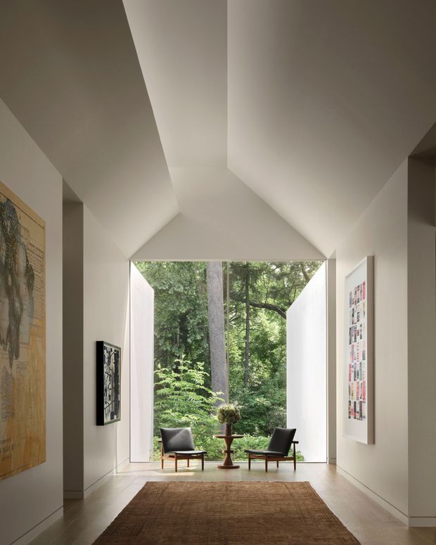 A refined, minimalist interior passage or gallery opens through a large floor-to-ceiling glazed opening toward a lush forest. Sculptural white angular ceiling planes create a dramatic vaulted effect, while two mid-century-style lounge chairs and a small pedestal table sit at the threshold. Pale walls and flooring, framed artwork, and a natural woven rug create a calm, gallery-like atmosphere.