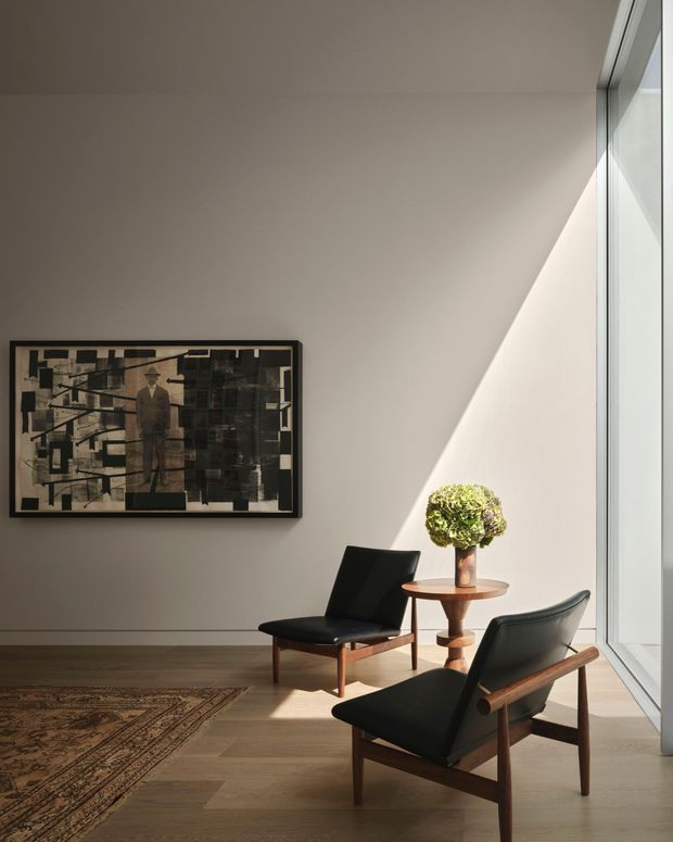 A refined, sunlit sitting area with two black leather mid-century lounge chairs arranged around a small round timber side table. The room has warm timber flooring, a large patterned rug, a neutral plaster wall, framed abstract artwork, and tall floor-to-ceiling glazing that casts a strong diagonal beam of light. The restrained furniture, clean lines, and natural materials create a calm mid-century-inspired interior.