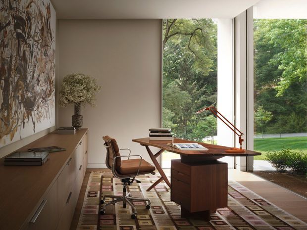 A refined home office with a mid-century-inspired timber desk, vintage-style swivel chair, low built-in storage cabinetry, patterned area rug, sculptural table lamp, and large floor-to-ceiling windows overlooking leafy gardens. A large abstract artwork provides a textured feature on the left wall. The room is minimalist yet warm, with strong natural light and a close connection to the landscape.