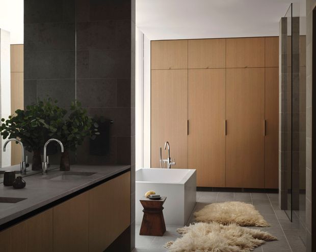 A refined contemporary bathroom with a freestanding white bathtub, wall-mounted timber cabinetry, dark stone-look tiled wall, grey tiled floor, glass shower enclosure, and a large stone vanity with curved metal basin tap. Warm wood cabinetry contrasts with dark tile surfaces, while layered wool rugs and greenery add softness.