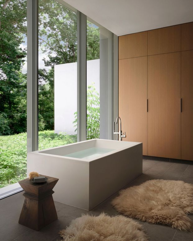 A serene contemporary bathroom with a freestanding rectangular bathtub positioned beside full-height windows overlooking lush greenery. The room features warm wood built-in cabinetry, slim dark window frames, large dark grey floor tiles, a minimalist chrome tap, and soft sheepskin rugs. Natural light and the garden outlook create a calm, luxurious atmosphere.