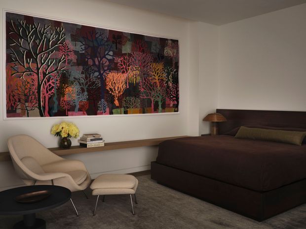 A calm, refined bedroom with a large colorful botanical artwork above a long built-in timber shelf. The room features a dark upholstered bed with a simple headboard, a neutral sculptural lounge chair and ottoman, a low black coffee table, a warm table lamp, and soft grey carpet. The restrained architectural shell allows the vivid artwork and dark furnishings to provide the main visual contrast.