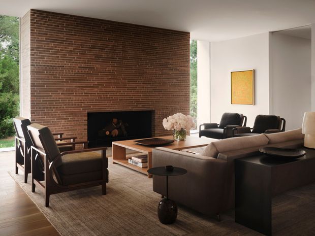 A refined modern living room with a large exposed red-brown brick fireplace wall, low white ceiling, expansive glazing to greenery, and a carefully arranged seating area. The room includes a large neutral sectional sofa, dark leather lounge chairs, a timber-and-wood coffee table, black side tables, a textured neutral rug, and understated artwork. The atmosphere is warm, calm, and sophisticated, with strong mid-century influences in the furniture and detailing.