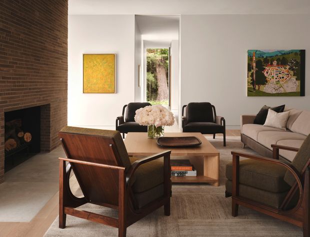 A refined living room with a mid-century-inspired seating arrangement around a low timber coffee table. The room features a full-height exposed red-brown brick fireplace wall with a dark firebox, pale smooth walls, timber-framed lounge chairs, a neutral upholstered sofa, a textured rug, floral arrangement, and framed artwork. A narrow glazed opening at the rear provides a view toward greenery.