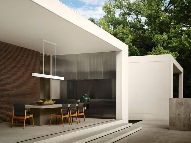 Contemporary open-plan dining and kitchen area opening onto a paved outdoor courtyard. The space features a long dining table with timber-framed chairs, dark built-in kitchen cabinetry, a brushed metal backsplash or wall surface, exposed brick, white geometric walls and roof planes, and a suspended linear pendant light. Broad concrete steps connect the dining area to the courtyard, with mature trees visible beyond.