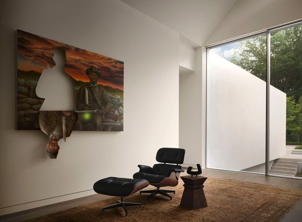 A refined minimalist living room with a black leather lounge chair and matching ottoman positioned beside a small sculptural side table. A large dramatic artwork hangs on the white wall, while expansive floor-to-ceiling glazing opens the room visually to a private courtyard and greenery. A large patterned rug adds warmth to the otherwise restrained interior.