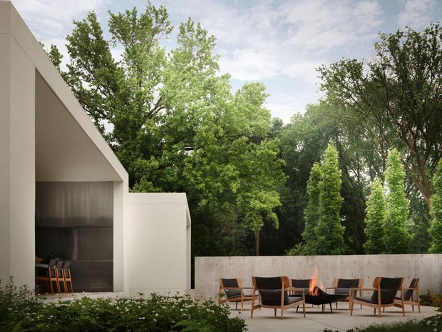 A contemporary outdoor entertaining terrace beside a minimalist pavilion, with a sharply angled roofline, pale rendered walls, a concrete privacy wall, and a built-in dark metal outdoor kitchen or barbecue zone. Mid-century-style timber lounge chairs surround a low fire pit, with dense mature greenery forming a private garden setting.