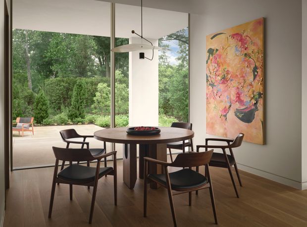 A refined dining area with a round dark-wood dining table and coordinating mid-century-style chairs, positioned beside expansive floor-to-ceiling glazing overlooking a lush garden. A large abstract artwork adds a colourful focal point, while a sculptural pendant light hangs above the table. The room has warm timber flooring, pale walls, and a calm connection to the outdoors.