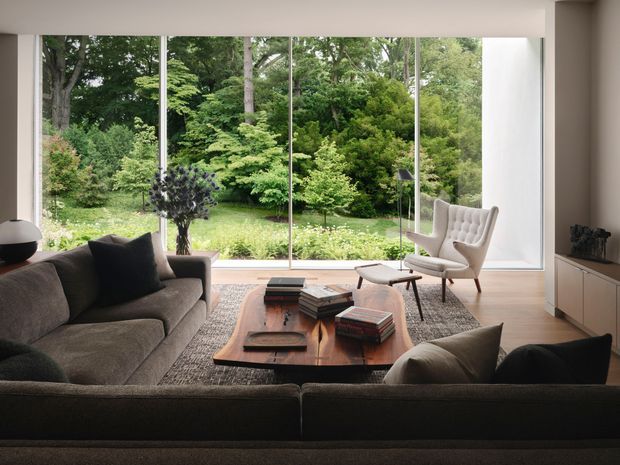 A refined living room with a large floor-to-ceiling glazed wall overlooking a lush garden. The space features a dark sectional sofa, sculptural lounge chair, low wooden coffee table with books, neutral textured rug, pale walls, and timber flooring. The composition has a strong connection to nature and a calm, mid-century-influenced contemporary interior.