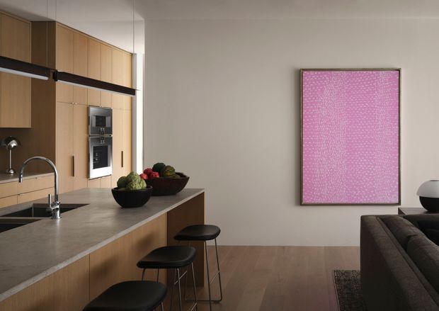 Contemporary open-plan kitchen and living area with warm timber cabinetry, a long stone island bench, integrated appliances, black pendant lighting and stools, and a dark sofa partially visible at right. A large pink artwork provides a bold accent against the light neutral wall, while timber flooring adds warmth.