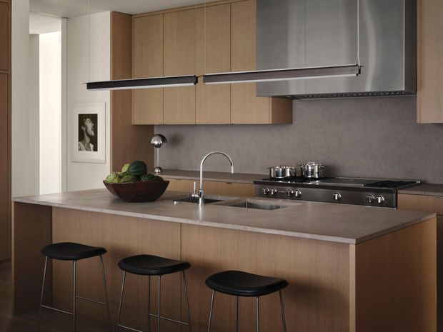 A contemporary kitchen with a large timber-clad island and three black stools, integrated sink and curved faucet, freestanding-style stainless steel range, and expansive timber cabinetry. A grey stone or composite benchtop and matching grey backsplash create a restrained, refined palette, complemented by a long linear pendant light and stainless steel rangehood.