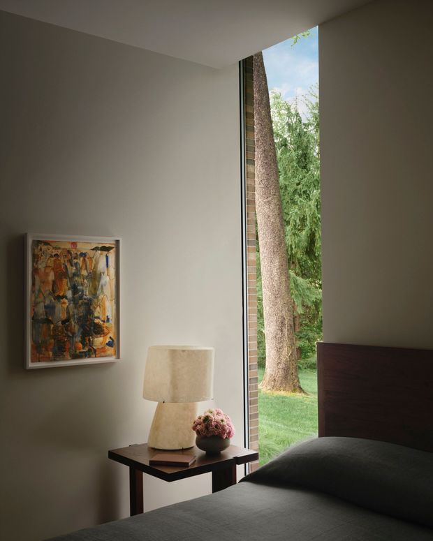 A refined bedroom interior with a dark upholstered bed and headboard positioned beside a tall, narrow floor-to-ceiling window. The window frames a mature tree and landscaped greenery outside. A dark timber side table holds a sculptural lamp, books, and a small floral arrangement, while an abstract artwork hangs on the soft neutral wall. The room has a calm, moody, minimalist character with strong natural light and a close connection to the garden.