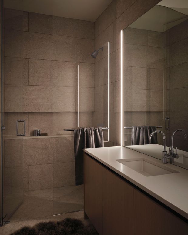 A moody contemporary bathroom with large warm taupe stone-look wall and floor tiles, a frameless glass walk-in shower, slim vertical integrated lighting, and a generous white double vanity with wood-toned cabinetry. A large mirror reflects the shower and curved chrome tapware, while dark towels and a soft rug add contrast.