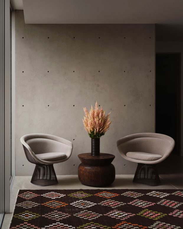 A moody contemporary interior vignette featuring two sculptural upholstered lounge chairs flanking a dark rounded wooden pedestal table with a vase of dried flowers. The chairs sit against a raw grey concrete feature wall with visible fixing marks, beside a dark doorway and a slim window or glazed opening. A richly patterned geometric textile rug fills the foreground. The composition has soft directional light, earthy neutral tones, and a refined minimalist arrangement.