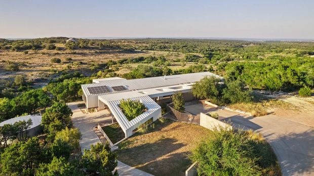 Aerial view of a low, sprawling contemporary residence set within a dry, semi-arid landscape. The building has pale metal pitched roofs, broad roof overhangs, extensive glazing, a covered entry or outdoor structure, and visible rooftop solar panels. Paved driveways and pathways connect the home to the surrounding native vegetation.