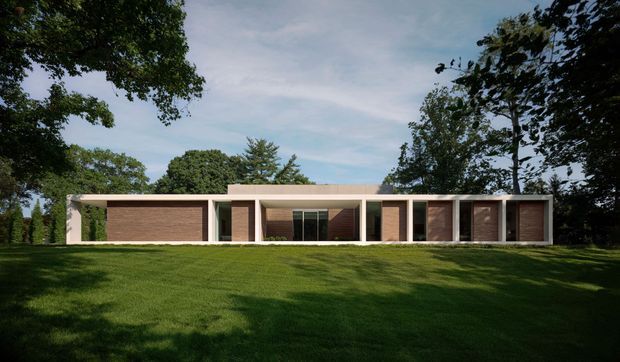 A long, low-slung single-storey residence set within a broad landscaped lawn and mature trees. The facade has a strongly horizontal composition with a flat roof, pale rendered or concrete structural walls, multiple recessed bays, dark timber-look wall panels, and large glazed openings. The restrained geometry and clean lines give the house a modern, minimalist appearance.