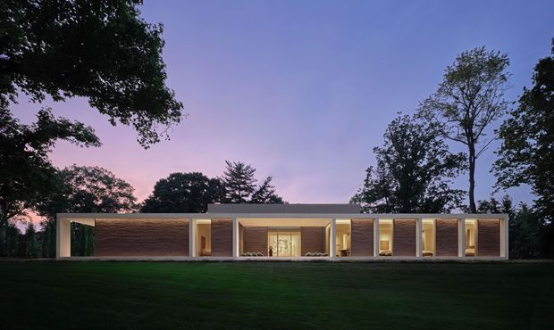 A long, low modern single-storey residence photographed at dusk, set within a dark landscaped lawn and mature trees. The facade is composed of repeated rectangular bays with warm illuminated glazing, pale structural or rendered wall planes, and dark horizontal timber-clad feature walls. The flat roofline and strongly horizontal composition give the house a restrained, minimalist contemporary appearance.