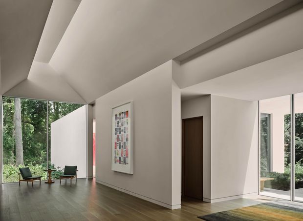 A bright, minimalist open-plan interior with a high, angular ceiling and exposed structural beams. Floor-to-ceiling glazing overlooks dense greenery, while two lounge chairs and a small table form a quiet sitting area. White plaster walls, dark timber flooring, a dark timber door, and a framed abstract artwork create a restrained contemporary setting.