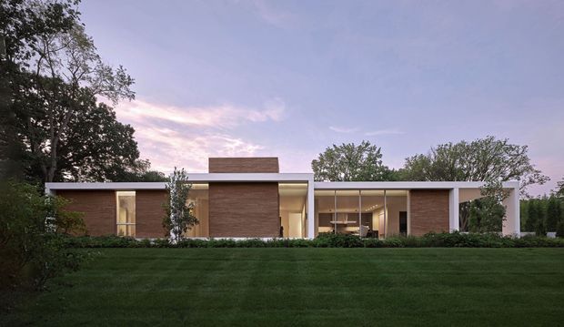 A wide exterior view of a low, single-storey modern residence set behind a manicured lawn and mature landscaping. The building has a strongly horizontal composition, flat rooflines, warm brown brick wall sections, white structural bands and columns, and extensive floor-to-ceiling glazing revealing a softly lit interior. The scene is photographed at dusk with a calm, refined atmosphere.