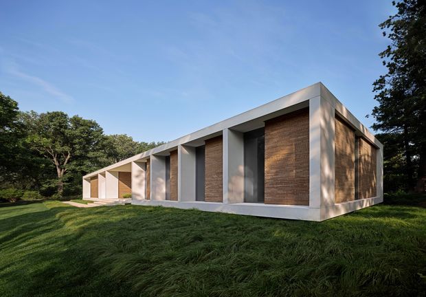 Exterior view of a low, single-storey modern house set within a wooded, landscaped site. The building has a strong horizontal composition, flat roof, deep recessed openings, pale rendered or concrete frames, and warm textured vertical brick feature walls. Long grass and mature trees surround the structure.