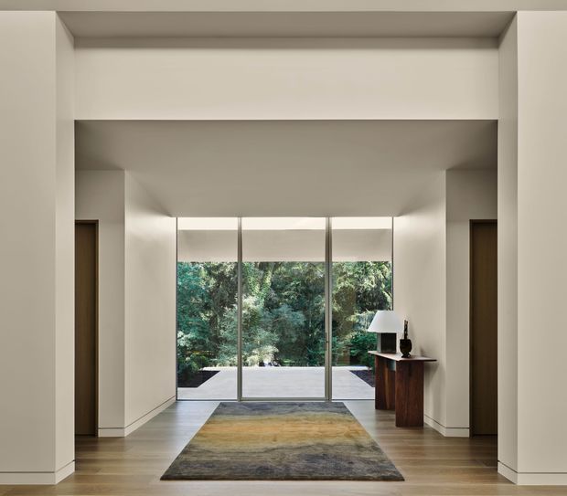 A refined, minimalist interior hallway or entry gallery framed by broad white plastered walls and a recessed ceiling. Warm timber flooring leads toward full-height glazed doors overlooking dense greenery and a paved outdoor area. A dark wood console with a table lamp and decorative object sits to the right, while a large muted abstract rug defines the central circulation space. Timber doors are visible on both sides, creating a symmetrical, calm composition.