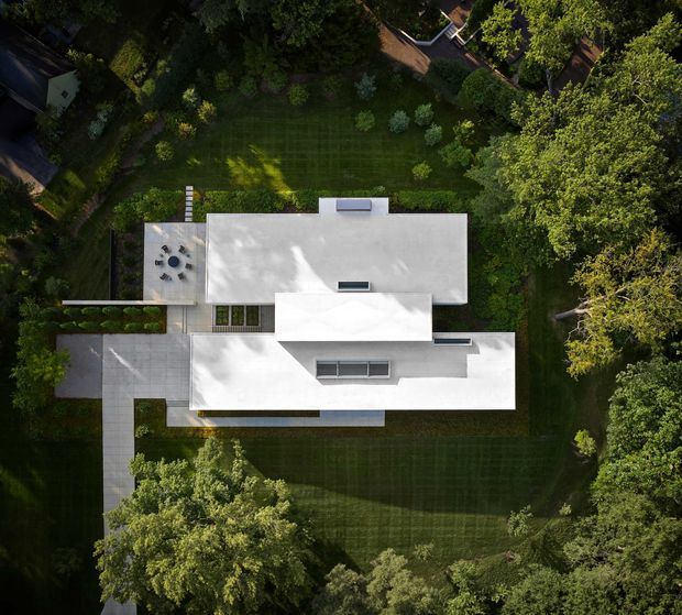 Aerial top-down view of a modern flat-roofed residence set within a large, mature landscaped garden. The building has crisp white geometric volumes, extensive pale concrete or paved terraces and walkways, a rooftop or terrace dining area, and dark linear roof openings or skylights. Dense trees and carefully maintained lawn surround the house, creating a secluded suburban garden setting.