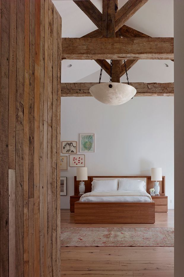 A warm, rustic bedroom viewed through a wide doorway, featuring a low solid-wood bed with white bedding, matching timber bedside tables, table lamps, framed artwork, and a large patterned rug. The room has prominent exposed, rough-hewn timber beams across the ceiling and a sculptural pendant light suspended from chains. Vertical reclaimed timber boards clad the wall and doorway on the left, contrasting with smooth white walls and wide timber floorboards.