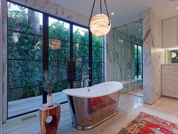 A luxurious bathroom featuring a freestanding polished metallic bathtub positioned beside full-height black-framed windows overlooking dense greenery. Veined marble-effect wall surfaces, a large frameless glass shower enclosure, white built-in vanity cabinetry, pale tiled flooring, a sculptural pendant light, and a patterned rug create a refined, spa-like interior.