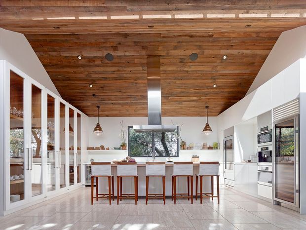 Bright, spacious contemporary kitchen with a long central island and seating for five, extensive white built-in cabinetry, integrated appliances, glass-front pantry or display storage, and a striking reclaimed timber-lined ceiling. Pendant lights and a stainless-steel rangehood add warmth and contrast, while large glazing brings in natural light and views of greenery.