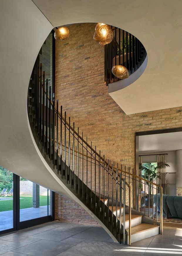 A dramatic interior view of a curved stairway winding around a tall exposed brick wall. The staircase has dark metal balusters, timber treads, and a sculptural sweeping plaster underside, with three warm pendant lights hanging in the stairwell. Large glazed doors open toward a landscaped exterior, while a living area is visible beyond the stair. The composition emphasizes the sculptural staircase, double-height circulation space, and contrast between brick, metal, timber, and plaster.
