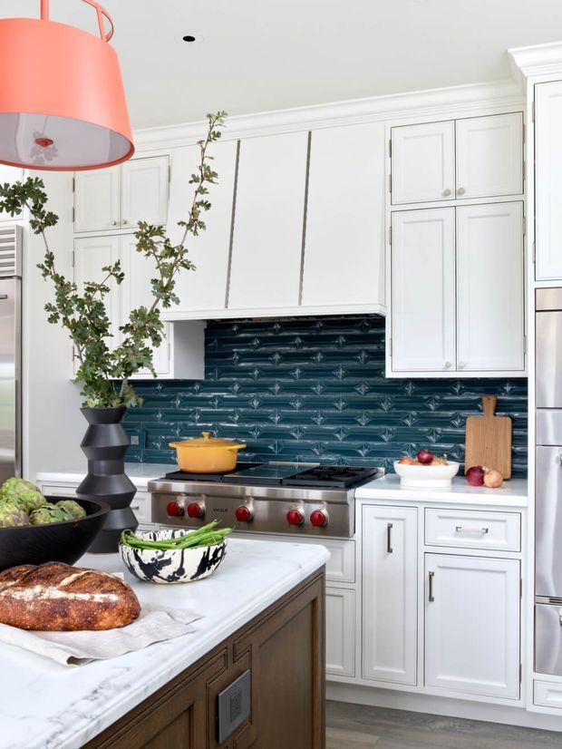 Bright, refined kitchen with white raised-panel cabinetry, a dark blue glossy textured tile backsplash, stainless-steel range and refrigerator, and a white marble-look island benchtop. The island includes dark timber cabinetry and is styled with bread, vegetables, cookware, and a sculptural hanging coral pendant light.
