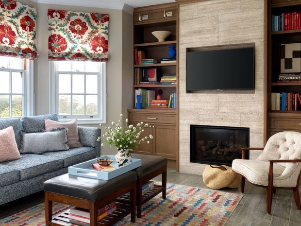 A refined living room with built-in dark wood shelving and cabinetry surrounding a television and fireplace faced in textured light stone. The room includes a grey sectional sofa, patterned upholstered armchair, dark wood coffee table and ottoman, colourful geometric rug, floral Roman shades, and a large window overlooking greenery. The overall look is warm, traditional, and comfortably layered.